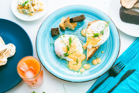 Poached eggs with hollandaise sauce on toast with crouton on a turquoise plate, next to a glass of orange juice and cutlery. High quality photoの写真素材