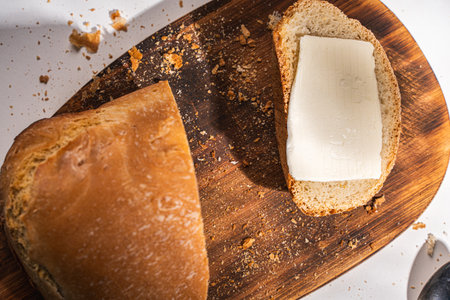 A slice of bread with butter and a cut loaf on a wooden board on a white table and background. High quality photoの写真素材