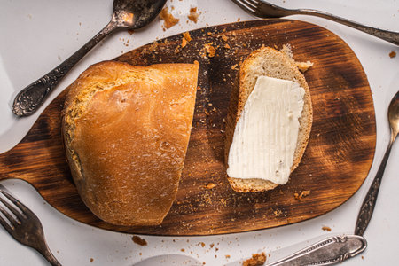 A slice of bread with butter and a cut loaf on a wooden board on a white table and background. High quality photoの写真素材