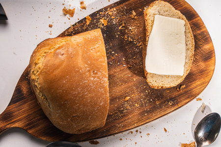 A slice of bread with butter and a cut loaf on a wooden board on a white table and background. High quality photoの写真素材