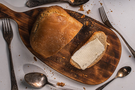 A slice of bread with butter and a cut loaf on a wooden board on a white table and background. High quality photoの写真素材
