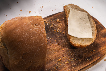 A slice of bread with butter and a cut loaf on a wooden board on a white table and background. High...