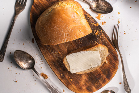 A slice of bread with butter and a cut loaf on a wooden board on a white table and background. High quality photoの写真素材