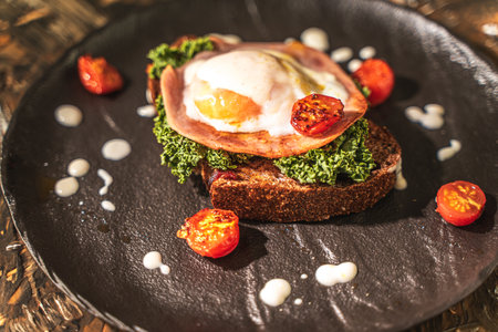 Toasted toast with bacon, poached egg, cherry tomatoes and lettuce on a black mica plate on a dark background. Close-up. High quality photoの写真素材