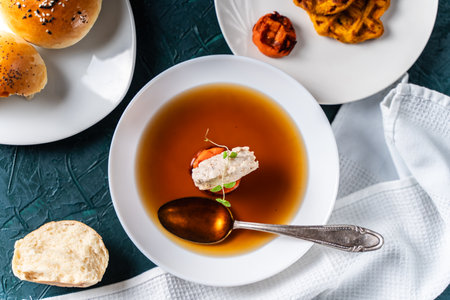 Soup or consomme, thick meat broth with carrots, chicken dumplings served in a white plate on a dark green background with sesame seed buns, napkin and spoon. View from above. High quality photoの写真素材
