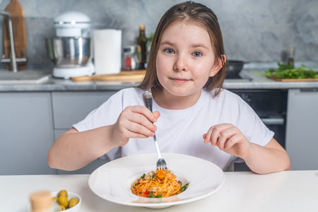 Cute little teen girl eating spaghetti on the background of gray kitchen at home sitting in white t-shirt at table. High quality photoの写真素材