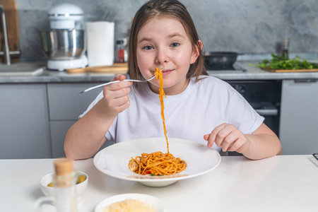Cute little teen girl eating spaghetti on the background of gray kitchen at home sitting in white t-shirt at table. High quality photoの写真素材