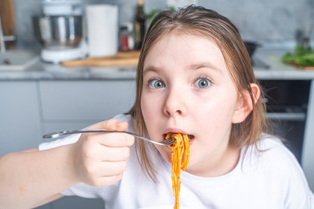 Cute, funny little teen girl eating spaghetti in the background of kitchen at home. Close-up. High quality photoの写真素材