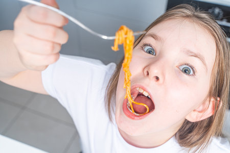Crazy little girl eating pasta, teen cheerfully putting pasta in her mouth in the background of the kitchen at home. Close-up. High quality photoの写真素材