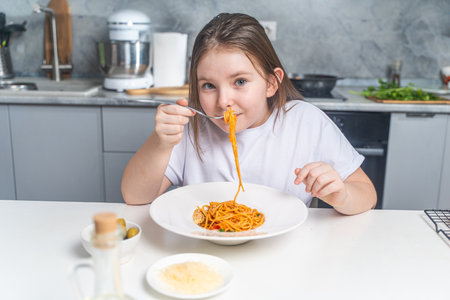 Cute little teen girl eating spaghetti on the background of gray kitchen at home sitting in white t-shirt at table. High quality photoの写真素材