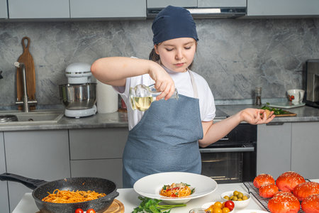 Little girl in apron cooking spaghetti in kitchen at home, pouring olive oil over cooked pasta in plate on white table. High quality photoの写真素材