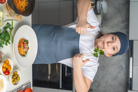 Portrait of a cute, little girl in the kitchen wearing an apron with cooked pasta, she gives a thumbs up and holds a sprig of parsley in her mouth. High quality photoの写真素材