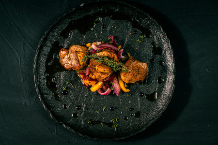 Slice of roasted pork tenderloin with vegetables, pink onion and yellow pepper on a black plate on a...