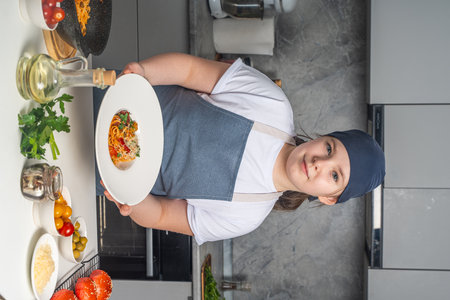 Portrait of a beautiful little girl in a gray bandana and apron holding a plate of pasta in her hands. The girl shows the spaghetti she has prepared. Close-up. High quality photoの写真素材