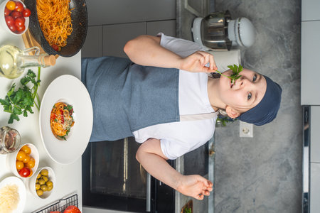 A pretty little teenage girl in a chef's apron is dreaming in the kitchen with a sprig of parsley in her mouth and a plate of spaghetti cooked by her on the table.の写真素材
