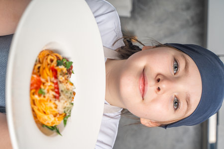 Portrait of a beautiful little girl in a gray bandana and apron holding a plate of pasta in her hands. The girl shows the spaghetti she has prepared. Close-up. High quality photoの写真素材