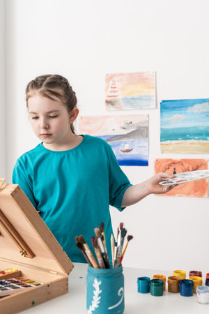A little girl against the background of the wall with her drawings paints a picture on an easel with a palette, brushes and jars of paint. High quality photoの写真素材