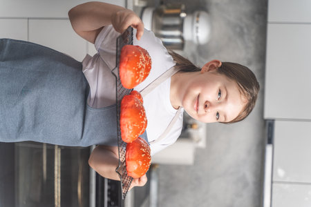 Cute little girl showing baked red sesame buns on the grill. Teenage girl in an apron against the background of the kitchen, at home Closeup. High quality photoの写真素材