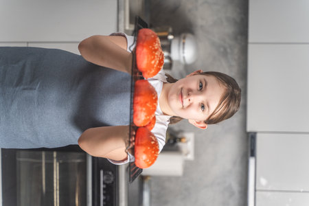 Cute little girl showing baked red sesame buns on the grill. Teenage girl in an apron against the background of the kitchen, at home Closeup. High quality photoの写真素材