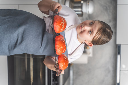 A little girl holds a tray of red sesame buns she baked. Girl in kitchen at home wearing white t-shirt and apron. High quality photoの写真素材