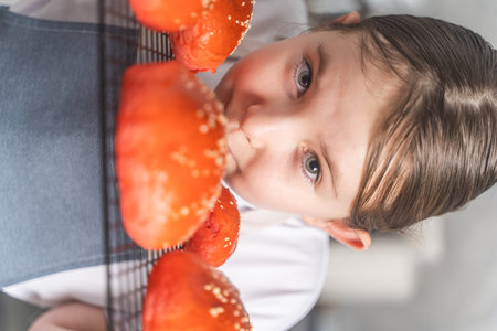 Cute little girl showing baked red sesame buns on the grill. Teenage girl in an apron against the...