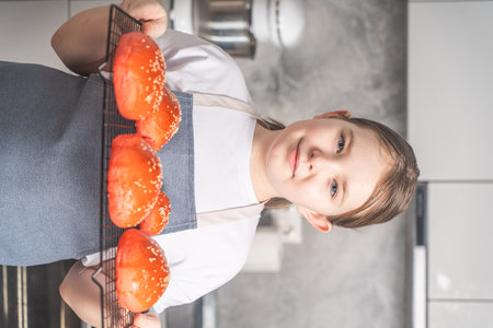 Cute little girl showing baked red sesame buns on the grill. Girl in an apron against the background of the kitchen, at home Closeup. High quality photoの写真素材