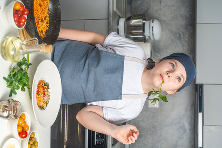 A pretty little teenage girl in a chef's apron is dreaming in the kitchen with a sprig of parsley in her mouth and a plate of spaghetti cooked by her on the table.の写真素材