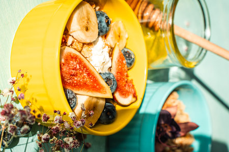 Granola with fresh banana, figs and blueberries on a light table with honey and flowers in a yellow and blue bowl. The concept of a delicious and healthy breakfast, a good start to the day. High quality photoの写真素材