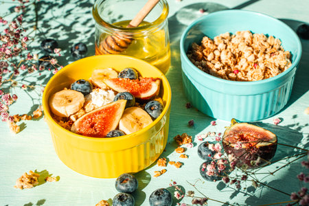 Granola with fresh banana, figs and blueberries on a light table with honey and flowers in a yellow and blue bowl. The concept of a delicious and healthy breakfast, a good start to the day. High quality photoの写真素材