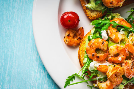 Toast with shrimp, arugula and avocado on a white plate with cherry tomatoes on a blue table. Space for text. Healthy and tasty breakfast concept. Top view. Flat lay. High quality photoの写真素材