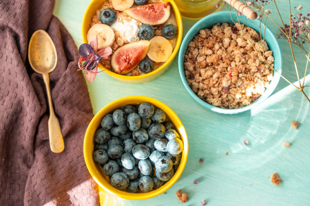 Granola and oatmeal with figs, blueberries and banana in a yellow bowl on a blue background are decorated with flowers. The concept of a healthy breakfast. High quality photoの写真素材