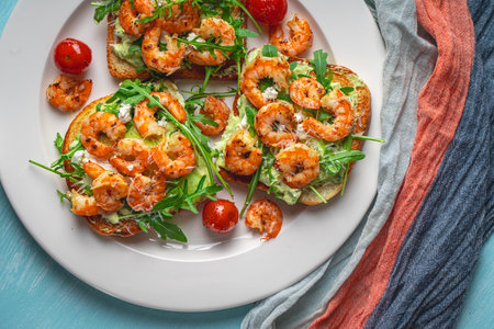 Toast or sandwiches with avocado oil, arugula and fried prawns on a white plate on a light blue background with a napkin. Healthy breakfast. View from above. Flat layout. Space for text. High quality photoの写真素材