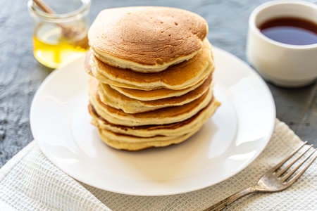 A pile of pancakes on a white plate with a napkin on a gray background with honey and a cup of tea. High quality photoの写真素材