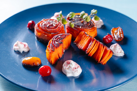 Carrot dessert, slices of carrots wrapped in a roll, fried and poured with honey with kumquat and cranberries on a blue plate. Healthy dessert. High quality photoの写真素材