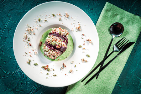 Beetroot with cottage cheese cream, nuts and pumpkin seeds on green spinach sauce served on a white plate with a napkin and cutlery. Restaurant serving of a healthy dish. Top view. High quality photoの写真素材