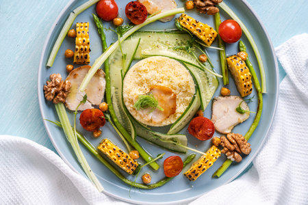 Couscous with chicken pieces, cucumbers, asparagus and corn, tomatoes and walnuts on a blue plate on a light background. View from above. Space for text. High quality photoの写真素材