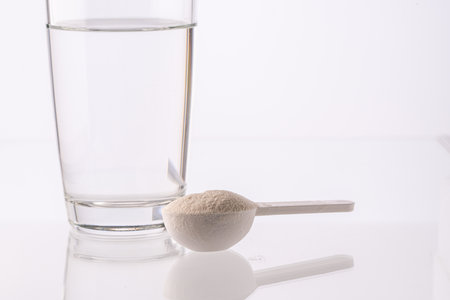 Collagen powder in a measuring spoon and a glass of water on a white background with reflection. Medicinal powder prepared for ingestion or dissolution in water. High quality photoの写真素材