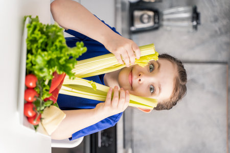 Beautiful little girl with celery in her hands and a plate of fresh vegetables. Girl in the kitchen at home on a plate of tomatoes, green salad, and peppers. High quality photoの写真素材