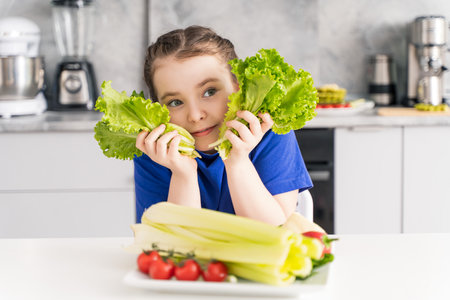 Cute little girl with lettuce leaves and a plate of vegetables with tomatoes and celery sitting at the table in the kitchen. Selected focus. High quality photoの写真素材