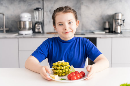 Cute girl sitting at the table with a healthy and delicious waffle breakfast with spinach, cheese and tomatoes. The concept of vitaminized nutrition for children. Selected focus. High quality photoの写真素材