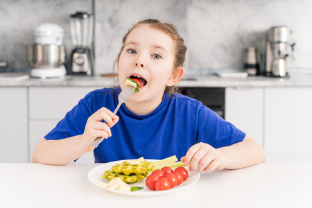 Happy little girl holding a fork in her mouth. A pre-teenage girl has breakfast in the kitchen at home with a plate of waffles, vegetables and cheese. Selected focus. High quality photo.の写真素材