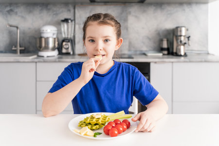 A delicious and healthy breakfast for a little girl. On the plate are spinach waffles, vegetables and cheese. The girl is sitting at the table against the background of the kitchen. Selected focus. High quality photoの写真素材