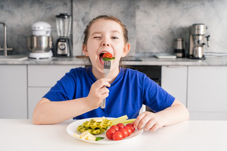Funny little girl holding tomato and cheese fork in her mouth. A young girl is having breakfast in the kitchen. Selected focus. The concept of a healthy breakfast. High quality photoの写真素材