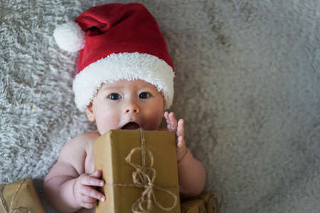 happy newborn baby in santa claus outfit making wow expression with eco friendly natural paper gift box for xmas lying on white backgroundの写真素材