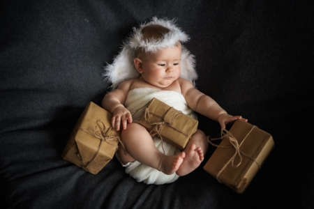 Cute little baby in angel costume with gift boxes on black background