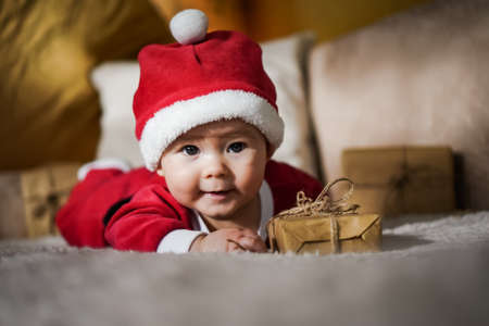 cute little newborn baby in christmas outfit with santa claus hat crawling on a couch in a cozy...