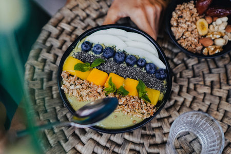 Bird view of a green vegan tropical fruit smoothie bowl with granola, mango, spirulina, coconut, nuts, chia seeds, mint on a wooden breakfast tableの写真素材
