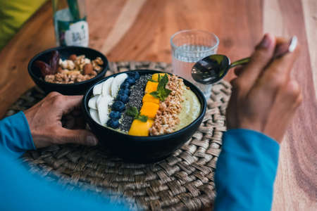 Bird view of a green vegan tropical fruit smoothie bowl with granola, mango, spirulina, coconut, nuts, chia seeds, mint on a wooden breakfast tableの写真素材