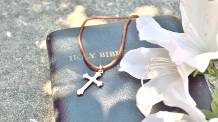 Silver and brown cross necklace, draped over a black, leather-bound Bible, with gold lettering, and azalea blooms to the right, all on a concrete sidewalk background.の写真素材