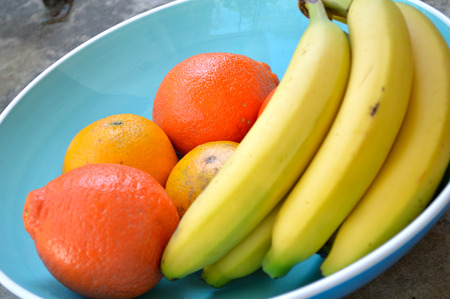Bananas, valencia oranges, and tangelos in a large turquoise blue bowl with white rim, sitting on a concrete surface in the sunlight. Fresh, ripe, tropical fruitの写真素材